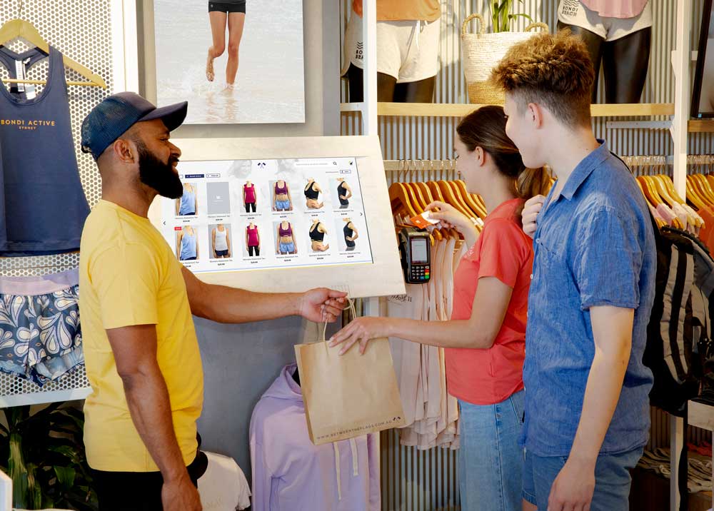 An employee hands a bag to custsomers who are looking at a touchscreen in a retail store